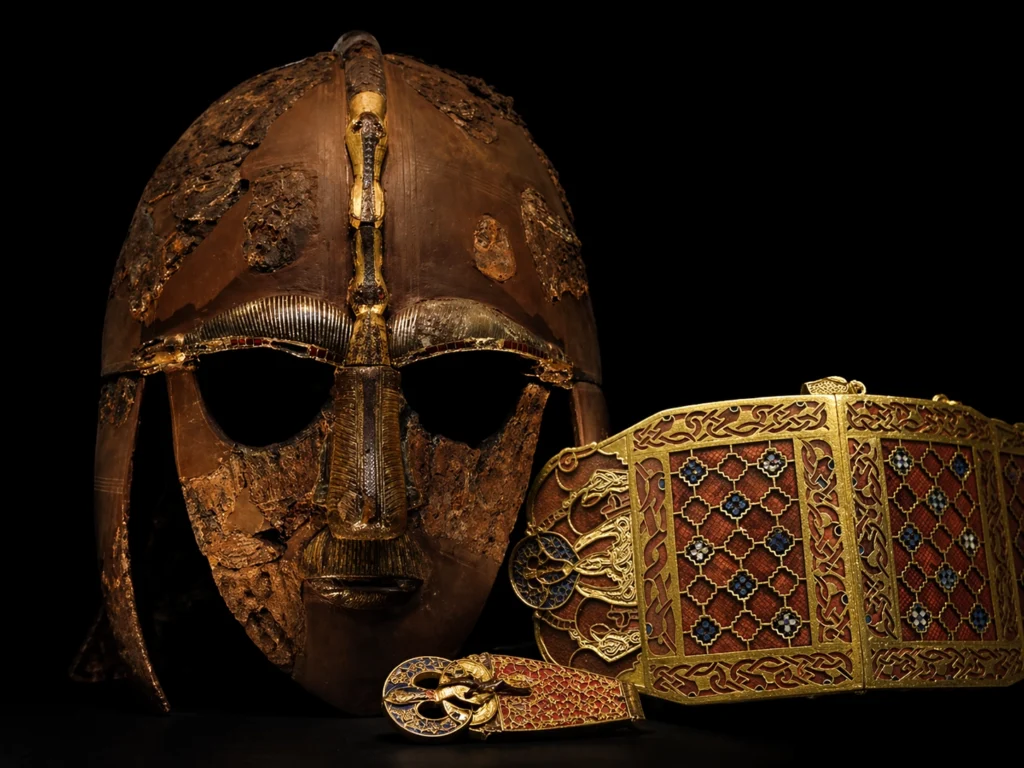 Dramatic image showing the Sutton Hoo helmet alongside richly decorated Anglo-Saxon treasures from early Britain against a black background.